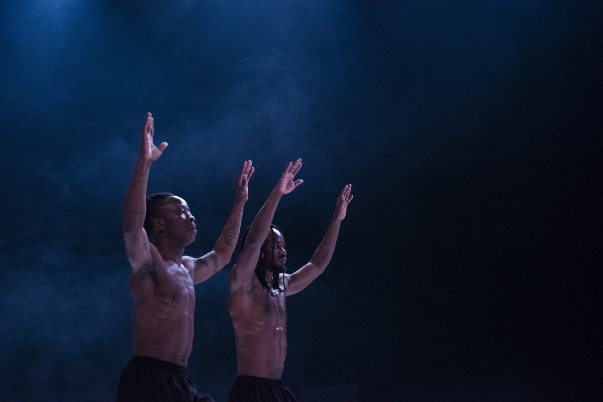 Two black men are standing together in a darkened room with arms outstretched.