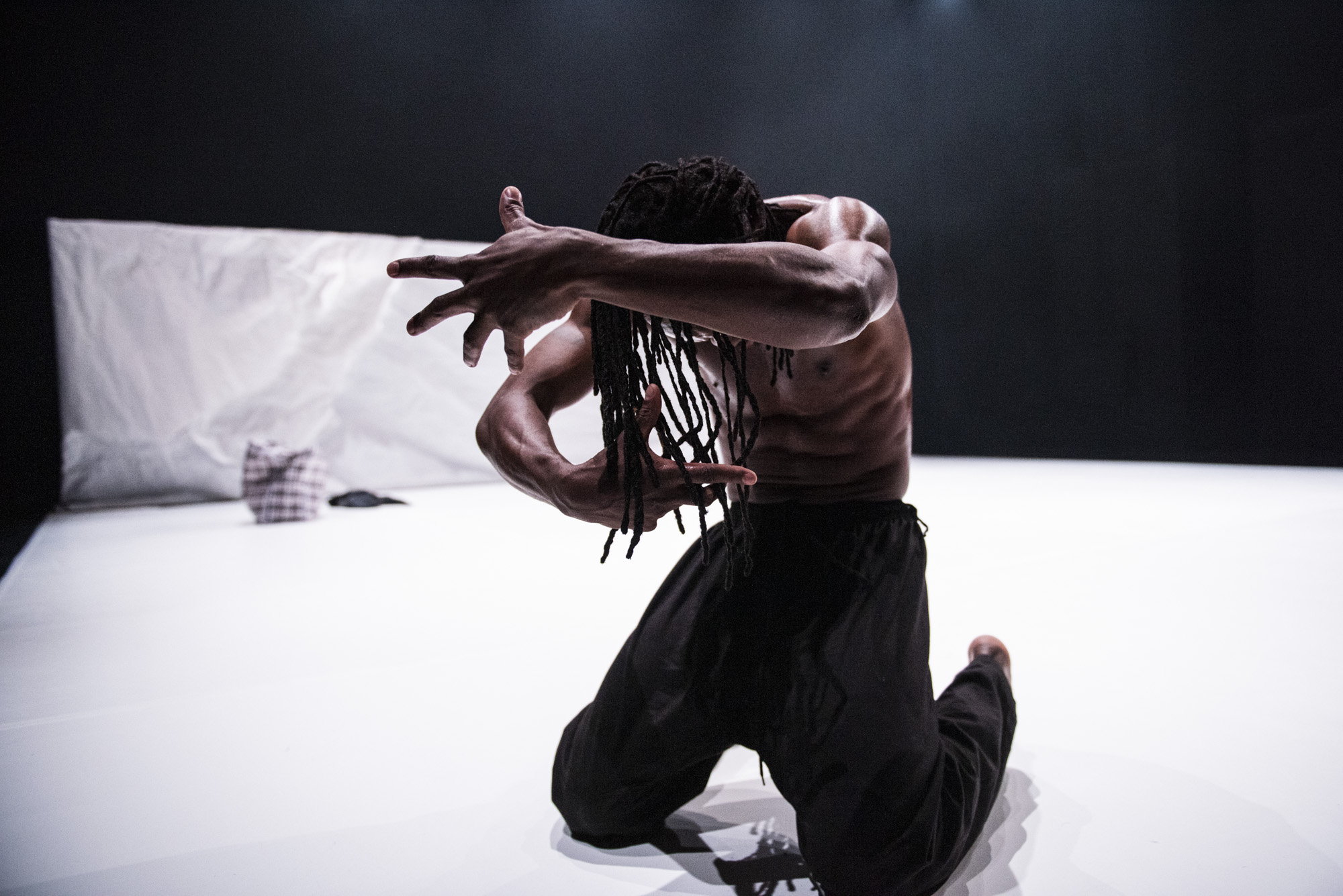 A black man on a white stage with a black backgroudn. He is kneeling with arms outstretched on either side of his head with his fingers splayed.