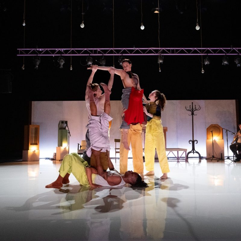 A group of 6 dancers on stage there is a black man lying on the floor, he is holding his partner who has their legs suspended in the air. To the left a man is holding his out a woman's extended arm. This woman is sitting on the shoulders of a hooded figure wearing a red cloak. There is a woman (dancer) standing on woman to the right holding the shoulder of the hooded figure and supporting the woman who is balanced on his shoulders