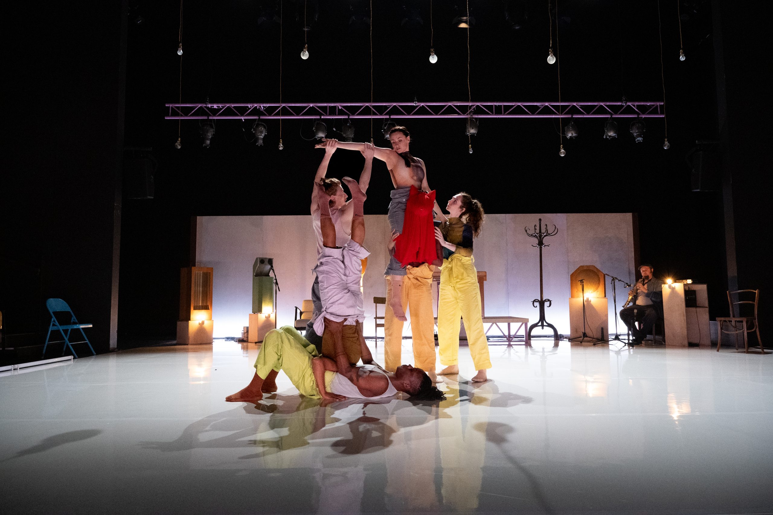 A group of 6 dancers on stage there is a black man lying on the floor, he is holding his partner who has their legs suspended in the air. To the left a man is holding his out a woman's extended arm. This woman is sitting on the shoulders of a hooded figure wearing a red cloak. There is a woman (dancer) standing on woman to the right holding the shoulder of the hooded figure and supporting the woman who is balanced on his shoulders